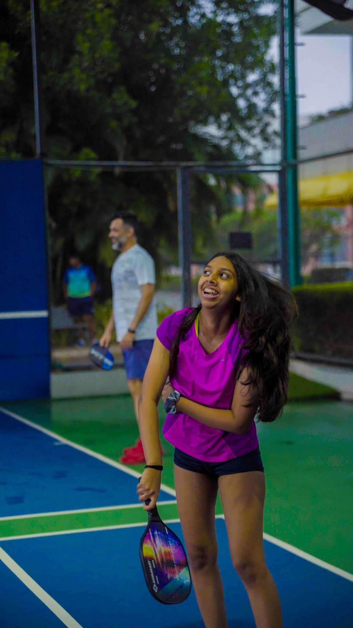 Young girl smiling and holding a pickleball paddle, playing on the court with an adult man.